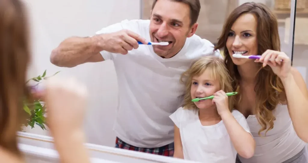 A family of three brushing their teeth together in a bathroom, demonstrating a simple daily dental health routine for maintaining healthy smiles